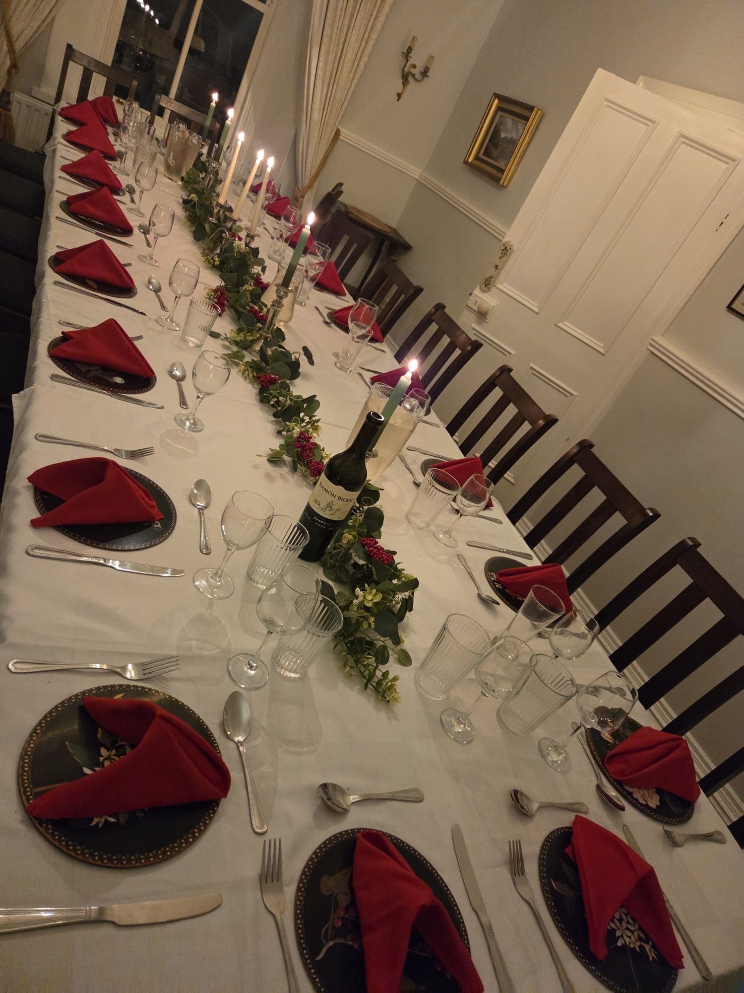 A long winter dinner table — white linen, red napkins, candles, and a eucalyptus and berry garland down the centre, set for a Christmas evening.