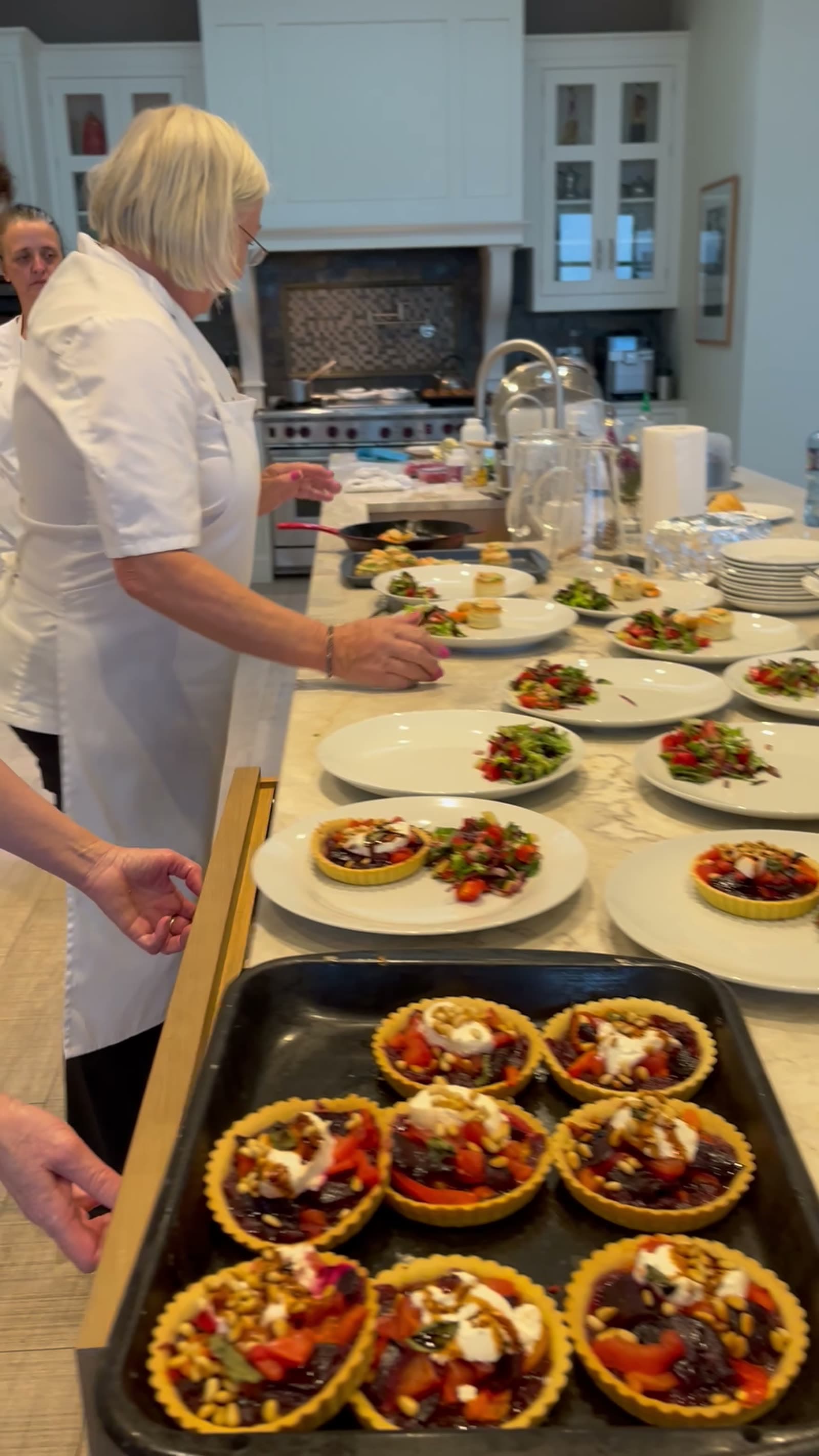Dóirín in chef whites at the marble kitchen island, plating a row of dressed-salad starters with a tray of beetroot-and-goat's-cheese tarts in the foreground; a second chef works on the next course behind her.