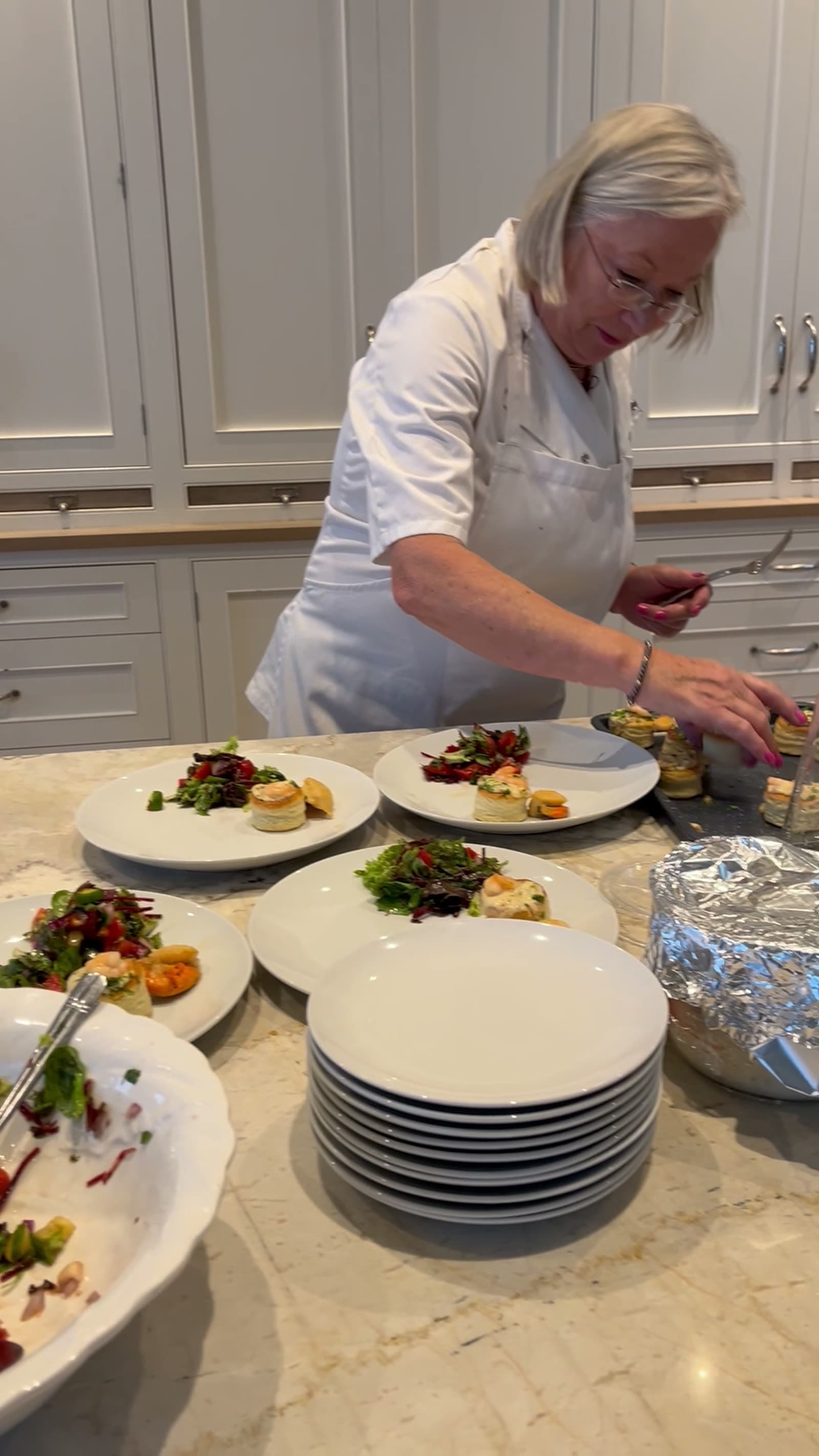 Dóirín plating vol-au-vents on dressed salad plates at the marble kitchen island, mid-service; a tall stack of clean white plates ready alongside and a foil-covered tray of warming food to one side.